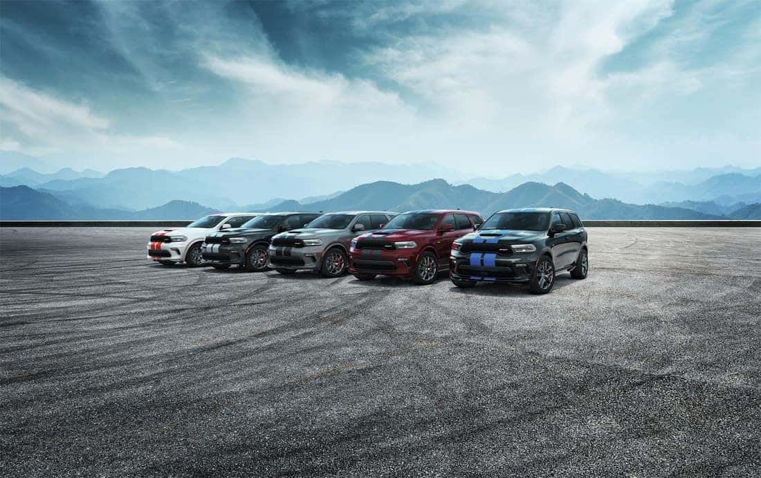 Family shot featuring 2022 Durango SRTs in the colors, from left to right: white with red stripes, black with silver stripes, gray with black stripes, red with gray stripes, and black with blue stripes parked with mountains in the background.