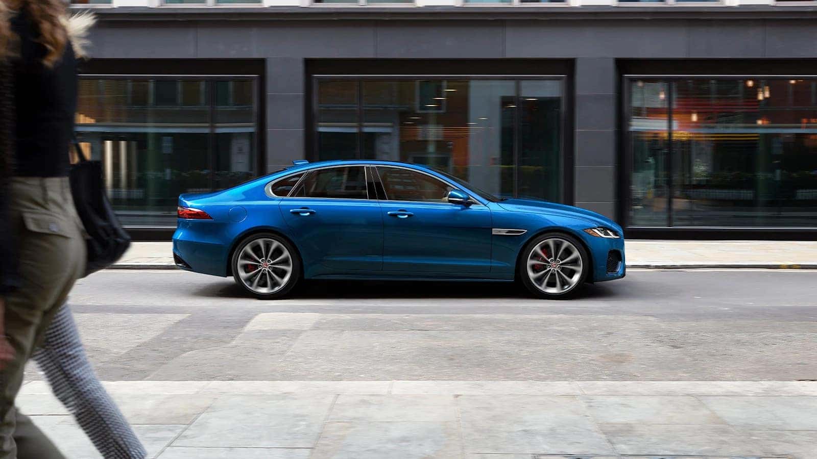 Blue Jaguar XF driving on the street in front of black buildings and a woman walking by 