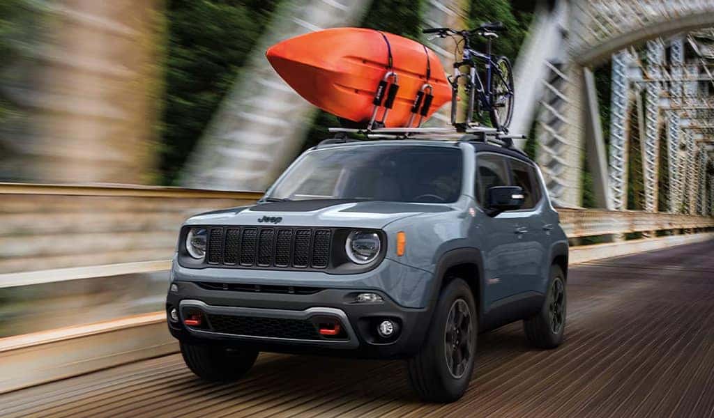 A silver 2023 Jeep Renegade driving on a bridge with a canoe and bike strapped to the top of the vehicle.