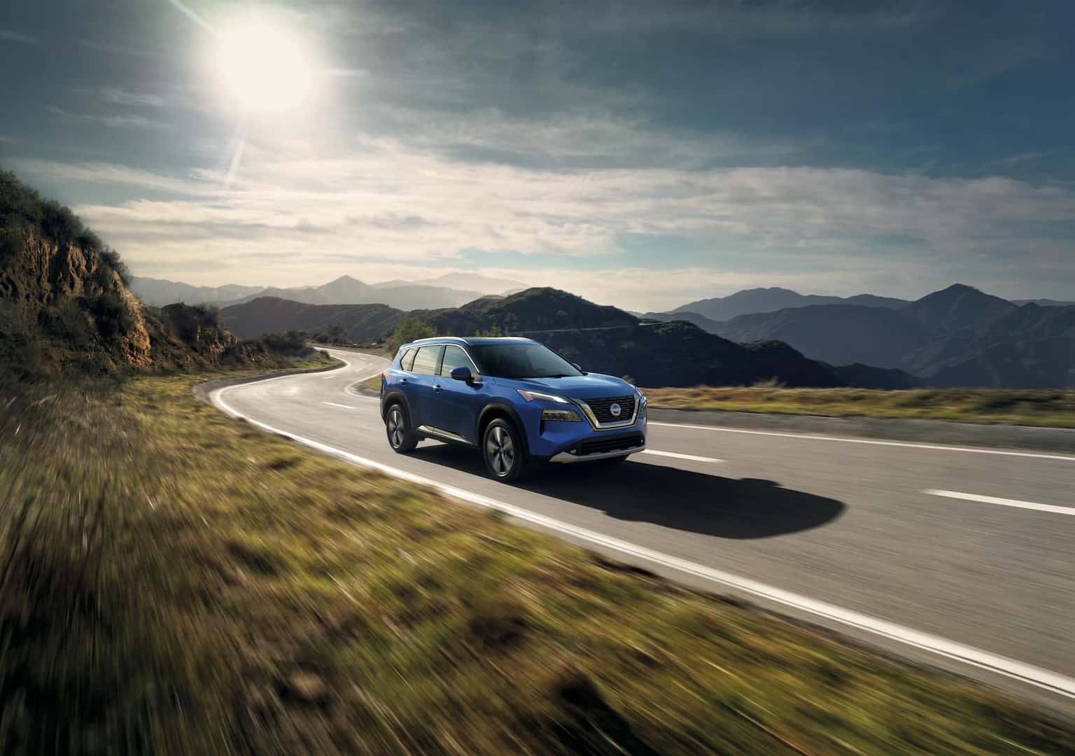 Blue 2023 Nissan Rogue driving on the road amongst mountains and grass