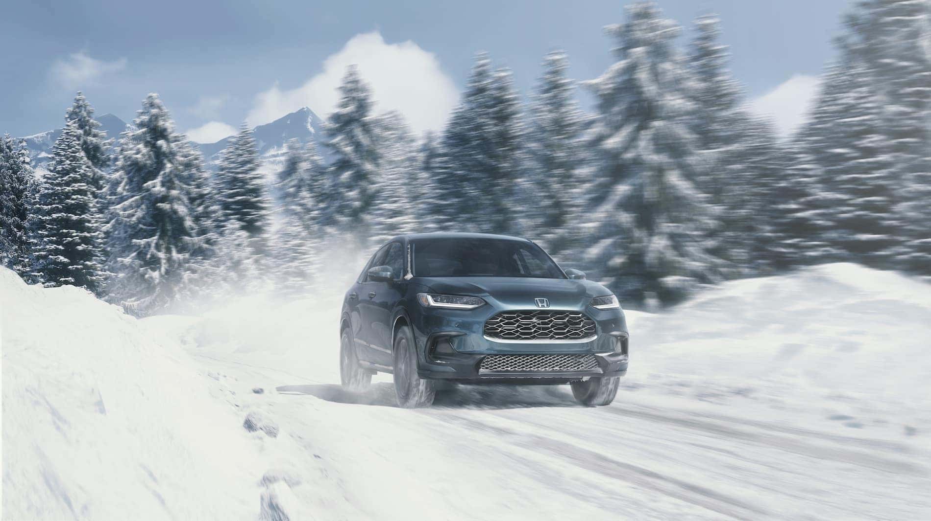 A blue 2023 Honda HR-V driving through snow with snow, trees, and mountains in the background.