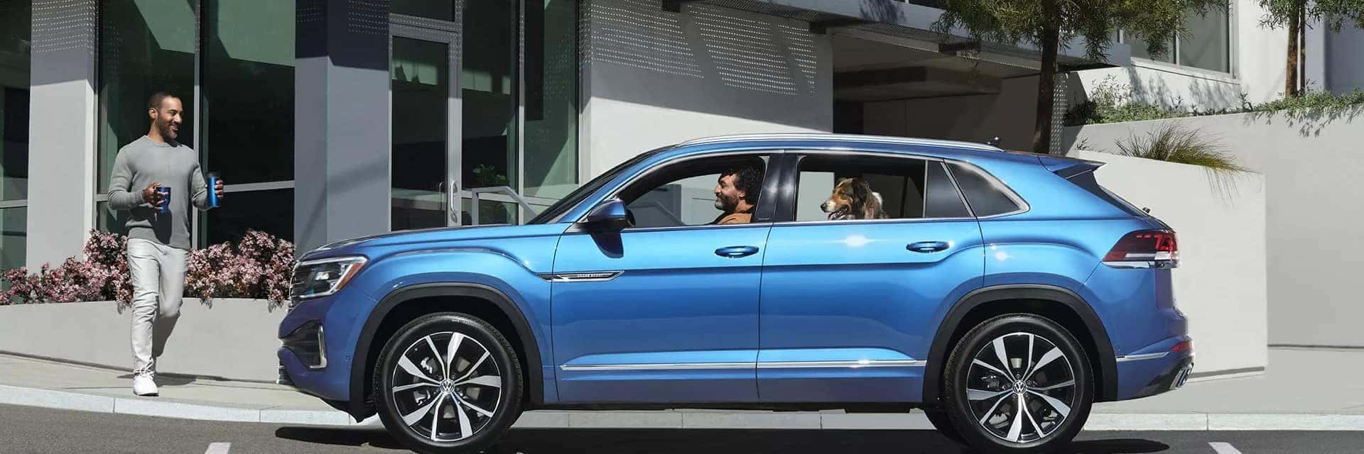 man driving blue VW Atlas Sport with dog in backseat picking up friend with coffee