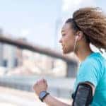 A young Black woman running outdoors in a city. She is wearing a blue shirt and an armband with her smartphone. She is wearing earbuds.