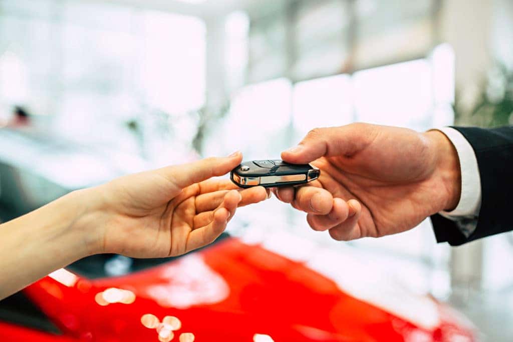 one hand giving another a car key fob in front of a blurred red car in background