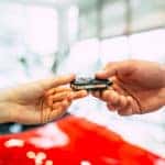 one hand giving another a car key fob in front of a blurred red car in background