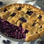 A blueberry pie with a slice missing. On a white and blue striped napkin with blueberries and serving utensils.