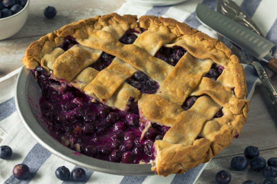 A blueberry pie with a slice missing. On a white and blue striped napkin with blueberries and serving utensils.