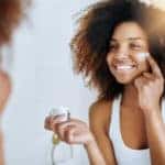A young woman applying moisturizer to her face in front of the mirror