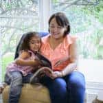 A young Latinx granddaughter sharing a chair with her grandmother and looking at a magazine together.