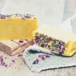 Two bars of yellow homemade soap with dried flower petals on the top of the soap bars. One bar is resting on a wooden block and the other is resting on a gray washcloth. Purple petals are scattered around the entire image.
