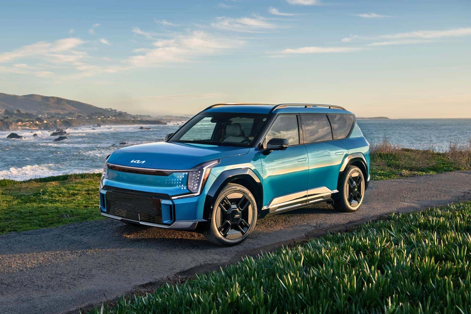 A 2024 KIA EV9 electric SUV parked on a scenic road with the ocean in the background, showcasing a serene coastal view.