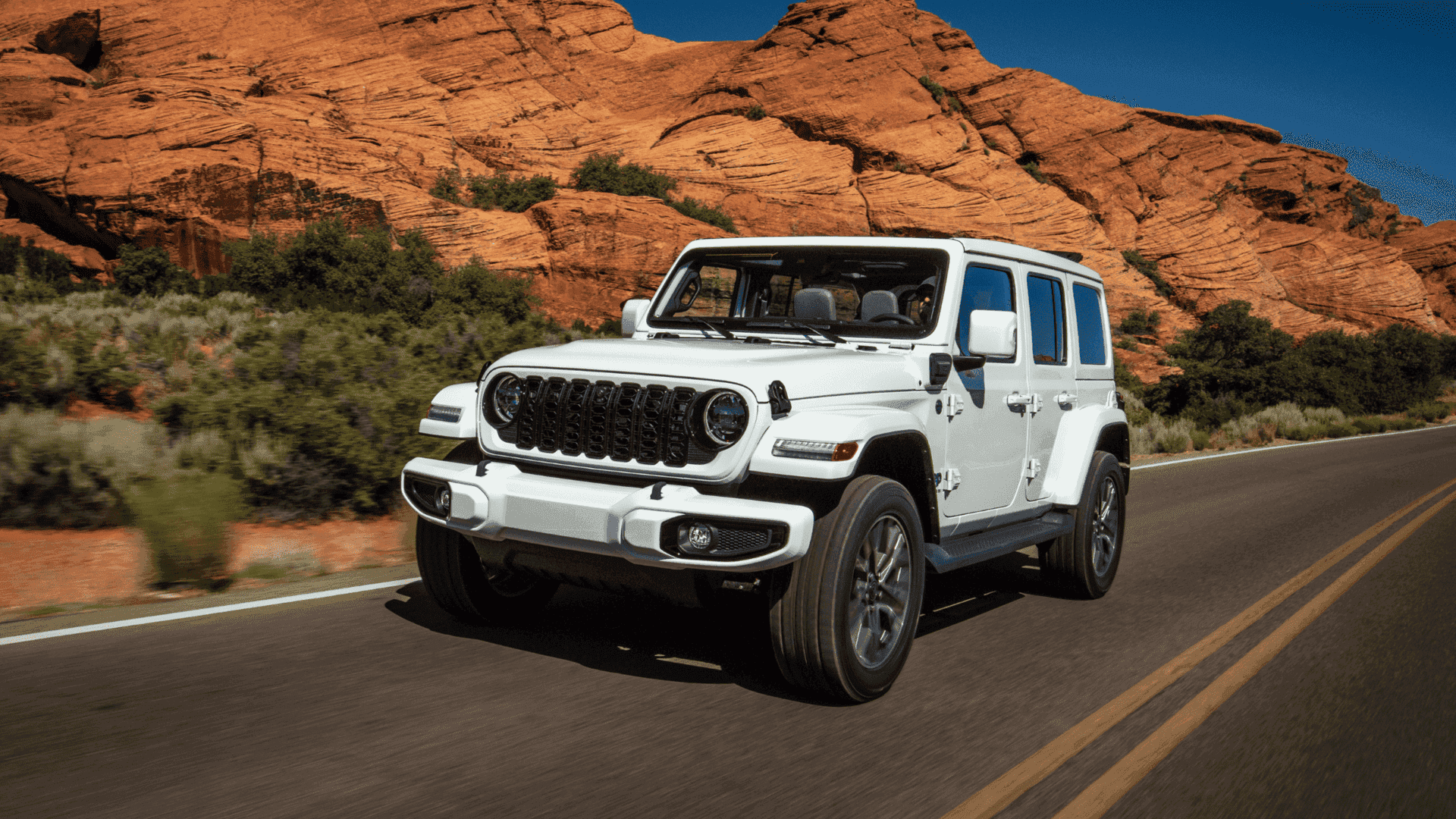 2025 Jeep Wrangler 4xe in white driving past red rock cliffs on a paved road