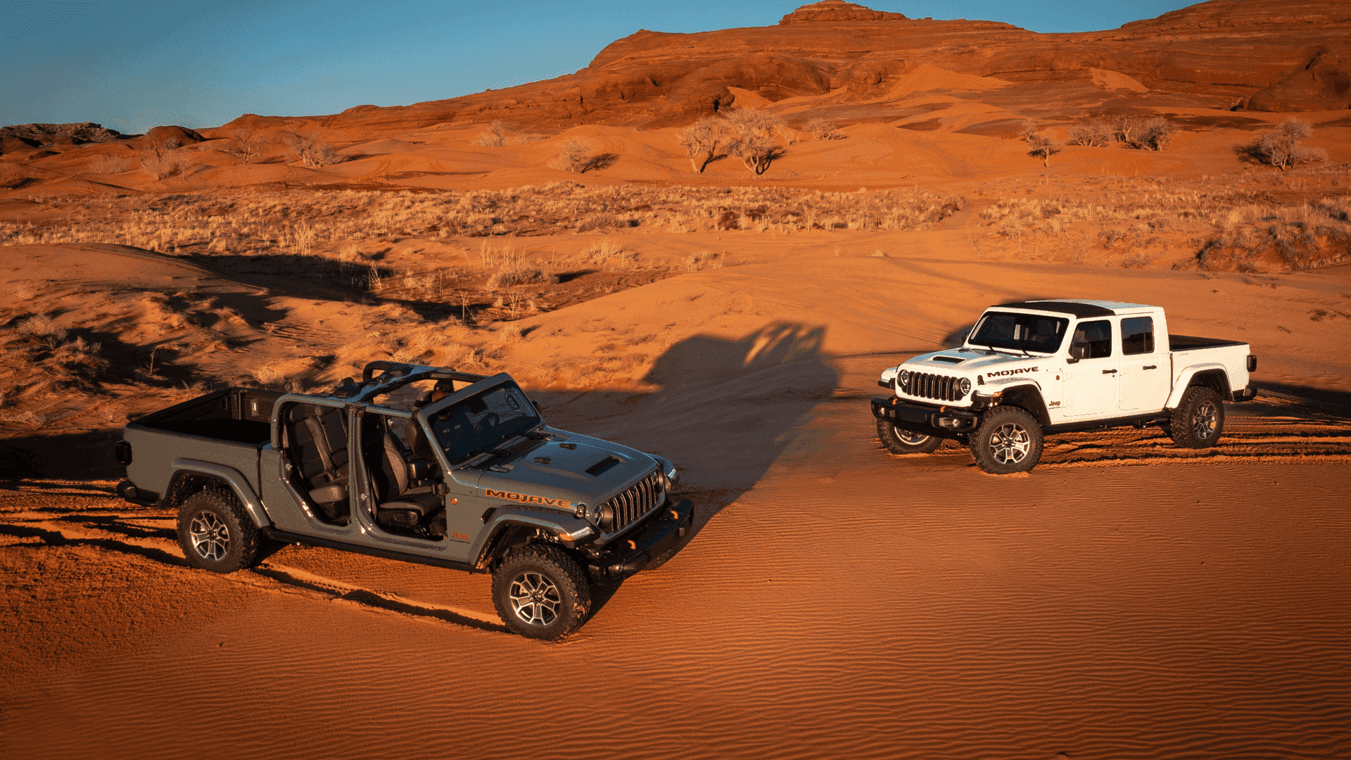 Two 2025 Jeep® Gladiator Mojave trucks, one with doors removed, parked on red desert terrain at sunset