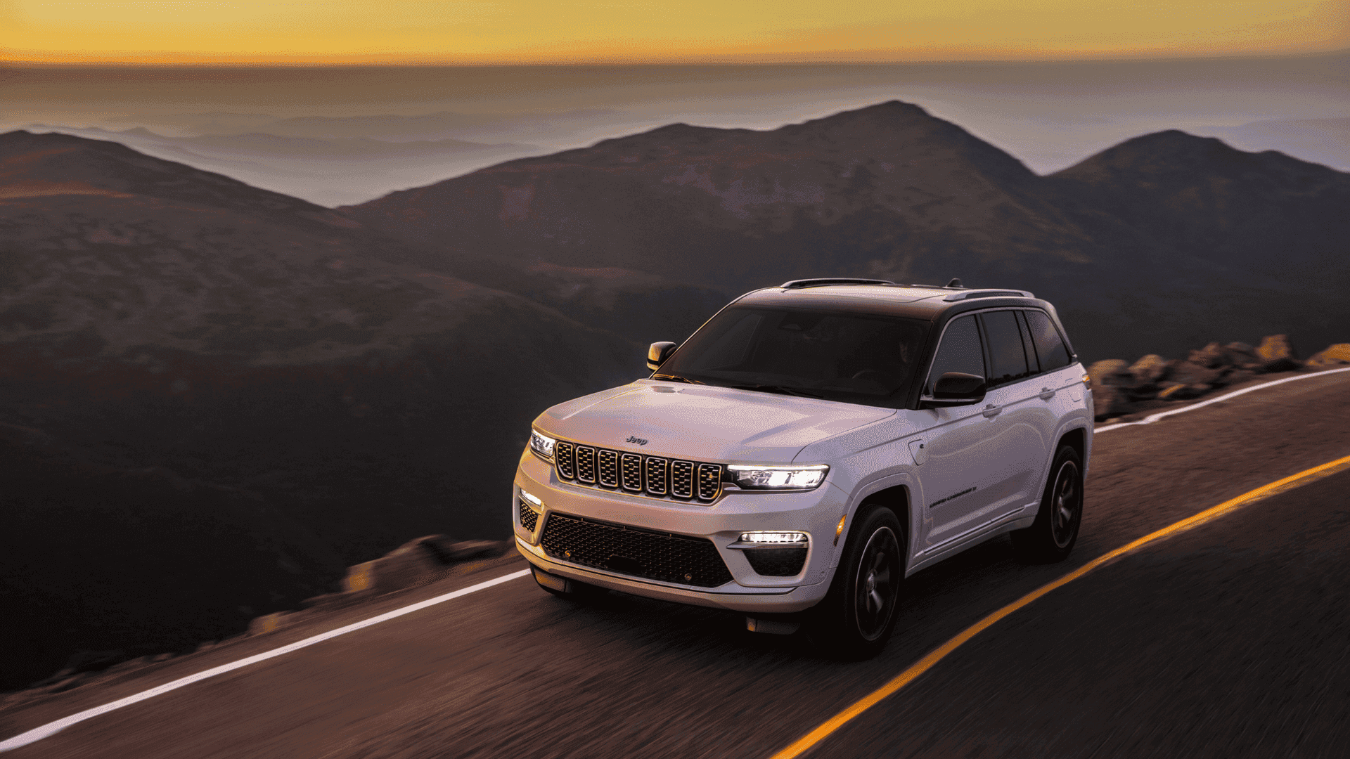 White 2025 Jeep® Grand Cherokee 4xe driving on a winding mountain road at sunset
