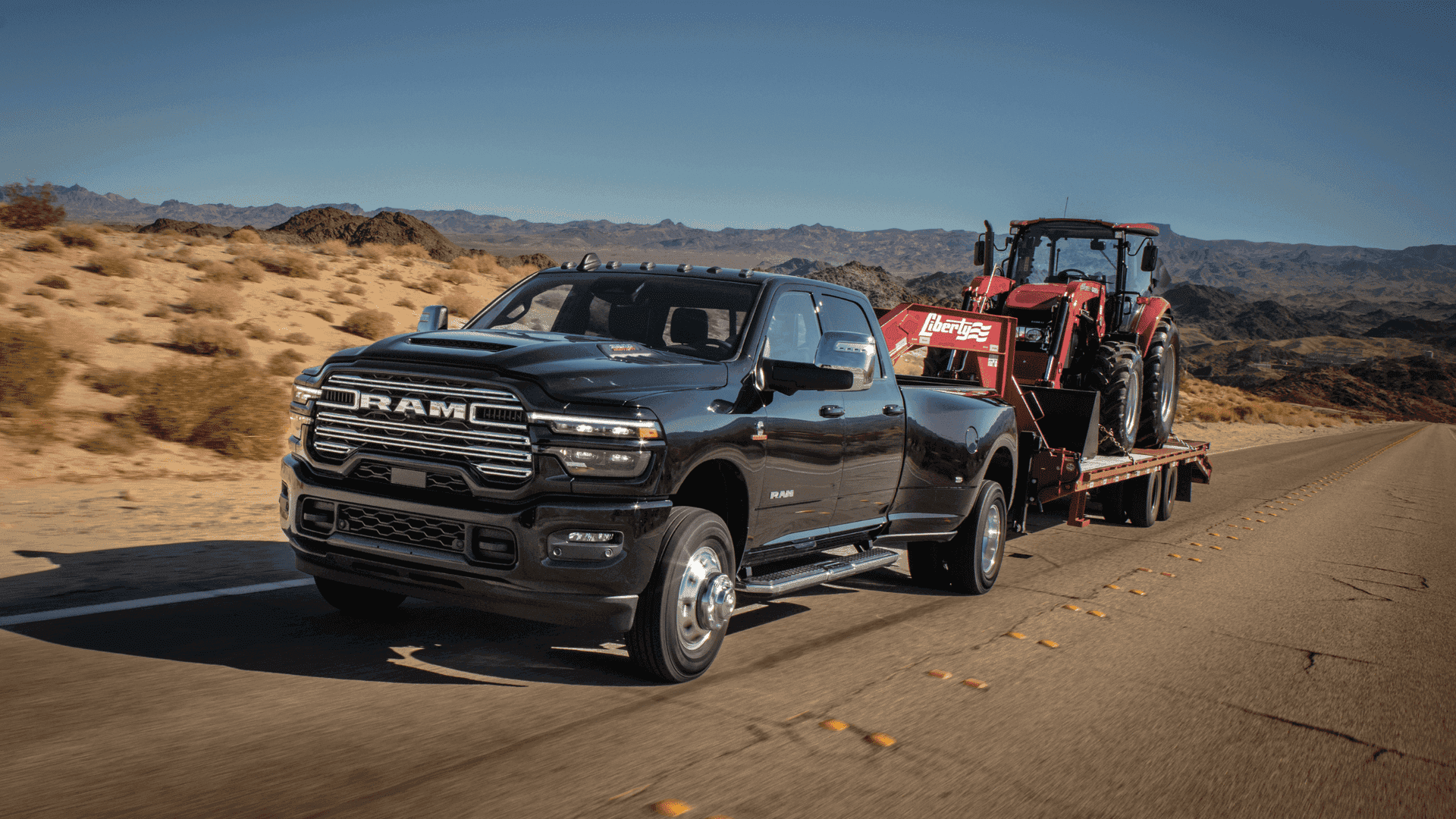 Black 2025 Ram 3500 towing red tractor on highway with desert mountains in the background