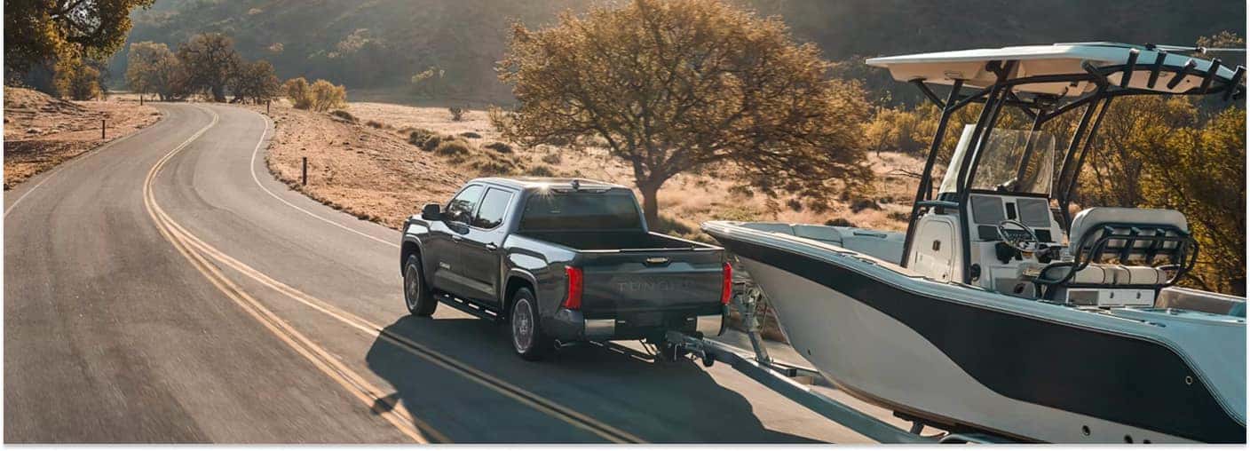 Dark gray 2026 Toyota Tundra towing a boat along a rural paved road.