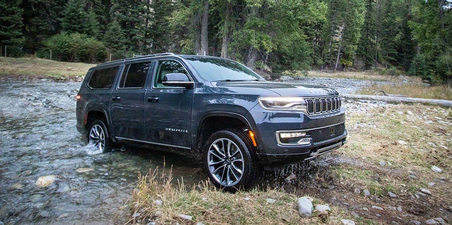 A navy blue Jeep Wagoneer driving out of a creek in a forest