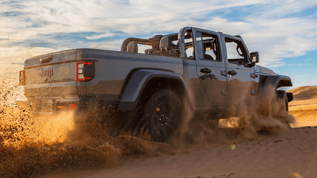 A rear view of a blue Jeep Gladiator kicking up sand as it drives through a desert