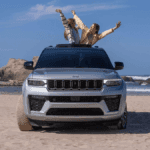 A silver Jeep Grand Cherokee on a beach with a pair of people popping out of the sunroof