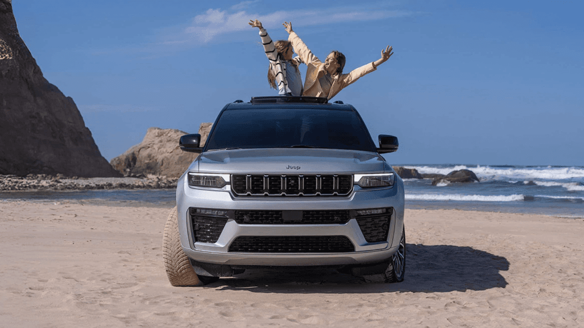 A silver Jeep Grand Cherokee on a beach with a pair of people popping out of the sunroof