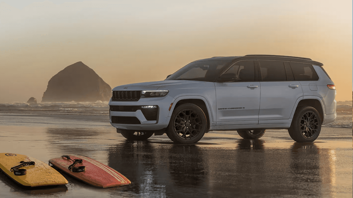 A white Jeep Grand Cherokee parked on a beach by the water near a yellow and red surfboard