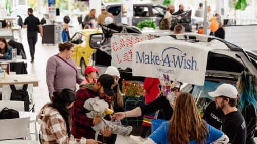 Families gather inside a bright car dealership showroom for a Make-A-Wish Minnesota event. Volunteers interact with children near decorated vehicles, including a yellow car and a white SUV with a Make-A-Wish banner displayed on its open trunk.