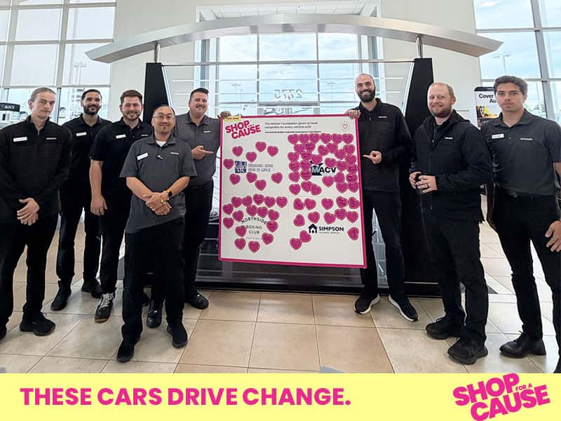 Walser employees stand behind a large pink Shop for a Cause board covered in signed paper hearts, featuring logos for CLUES, Northside Boxing Club, and Simpson Housing Services, in a dealership showroom.