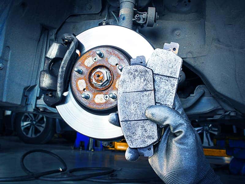 Close-up of a mechanic's gloved hand holding worn gray brake pads next to a rusty brake rotor during inspection..