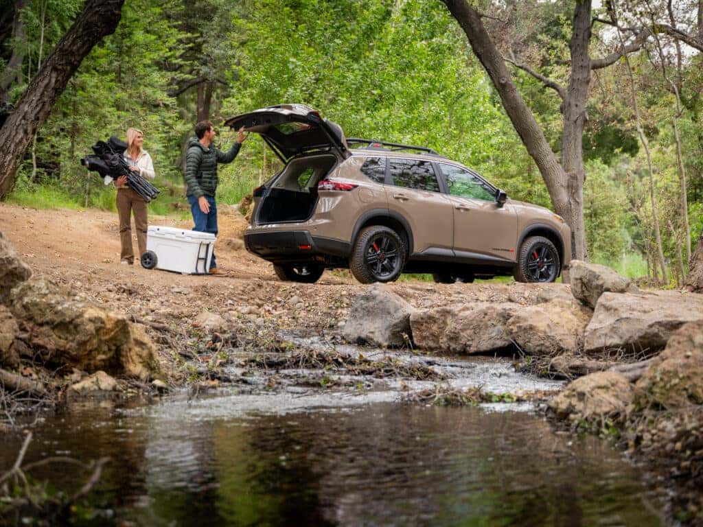 2025 Nissan Rogue Rock Creek edition parked next to a creek with two people unloading camping supplies